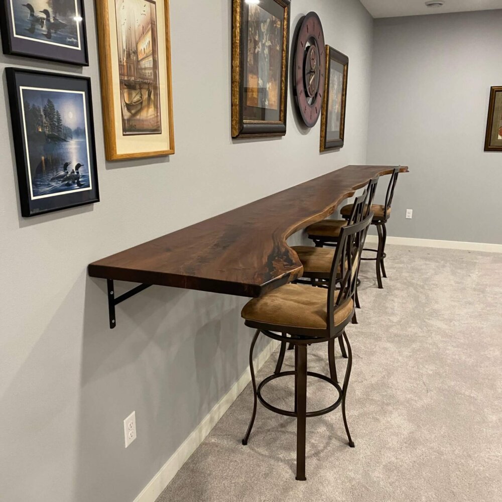 A minimalist room features a high wooden counter with a live edge, wall-mounted with black brackets. Three brown cushioned bar stools with ornate metal backs are placed along the counter. Reflecting the latest furniture trends, the walls are adorned with framed artwork and paintings.