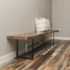 The Black Walnut Live Edge Bench, featuring a sleek seat crafted from a live edge Black Walnut slab and supported by black metal legs, sits against a grey wall in a corner. Two striped throw pillows rest on the bench. The floor consists of rustic, weathered wood planks showcasing an array of tones and textures.