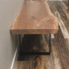 A close-up shot of the Black Walnut Live Edge Bench showcasing its rustic, natural design, positioned against a wall. The bench features a thick wooden slab top paired with sturdy black metal legs. The floor presents an intricate blend of dark and light wooden planks in diverse shades and textures.
