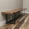 The Black Walnut Live Edge Bench, featuring its distinctive irregular edges and supported by sleek black metal legs, is positioned against a beige wall just above a floor vent. It rests on a wood-patterned floor exhibiting various shades of brown. The minimalist design enhances the space's rustic appeal.