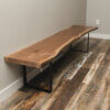 The Black Walnut Live Edge Bench, with its stunning long design and live-edge craftsmanship, is positioned against a light gray wall. It features two black metal legs and stands on a floor adorned with mixed-tone wooden planks. In the background, a white electrical outlet is visible on the wall.