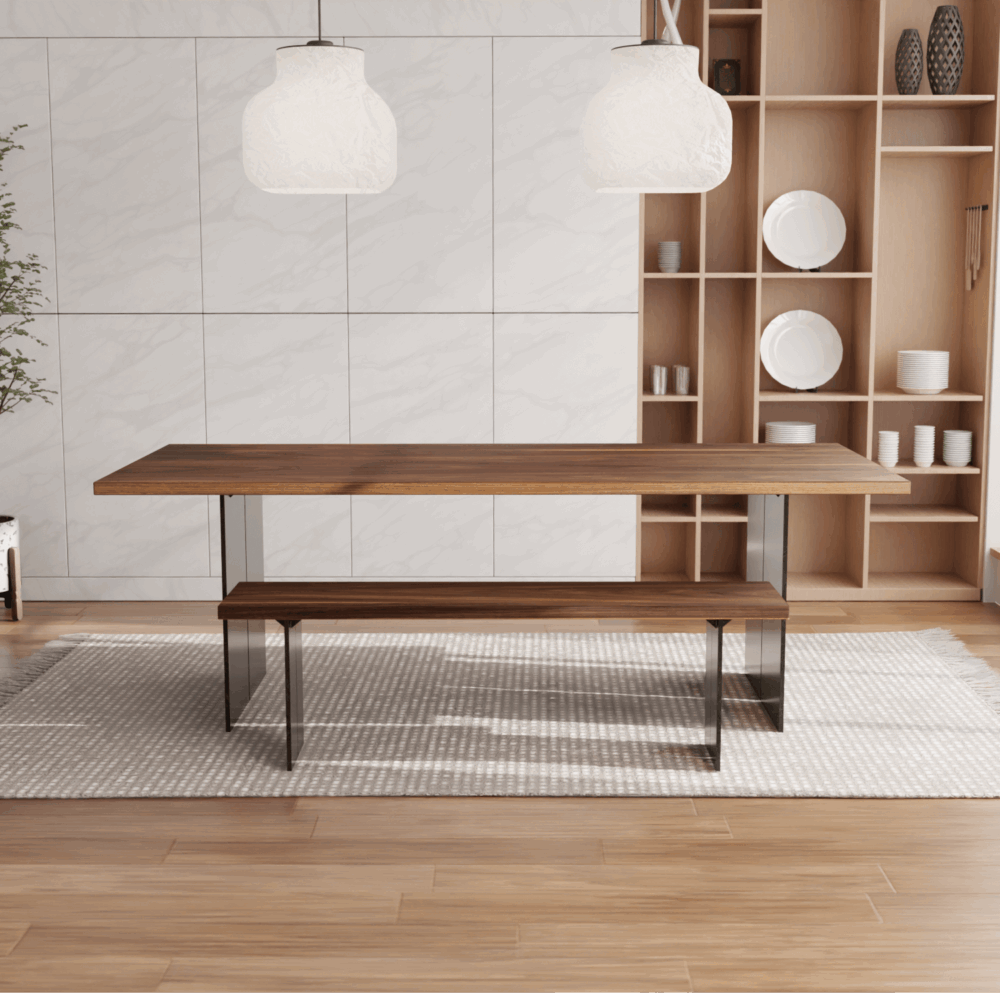 A minimalist dining room showcases The Steel Bennett Dining Table accompanied by its matching bench, both featuring sleek metal legs. Overhead, a pair of white pendant lights are suspended from the ceiling. In the background, shelves display neatly arranged dishes, while a potted plant is situated in the corner.