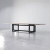 A sleek, minimalist dining table with a rectangular wooden tabletop and a black metal geometric frame, The Westbrook Conference Table perfectly balances modern style and functionality. Placed on a polished concrete floor against a plain, light-colored wall, this piece exemplifies the elegance of The Westbrook Conference Table.