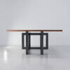 The Westbrook Square Dining Table features a modern, minimalist design with a sleek dark rectangular wooden top mounted on an artistic black metal base. The interconnected geometric shapes of the base provide a sculptural and contemporary aesthetic, perfectly complementing the table. This striking piece is set against a plain, light-colored background.