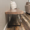 A Black Walnut Live Edge Bench with black metal legs rests against a beige wall on a polished wooden floor. The bench features a striped cushion, and a beige sofa is partially visible in the background to the right.