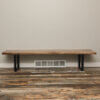 A Black Walnut Live Edge Bench with black metal legs sits on a wood-patterned floor against a plain beige wall. A white vent cover is visible on the wall beneath the bench.