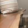 A close-up shot of the Black Walnut Live Edge Bench, accented by a light brown cushion with white stripes, set against a light gray wall. The rich texture and color of the bench are beautifully complemented by the wooden floor.