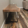 The Black Walnut Live Edge Bench, showcasing a beautiful live edge and supported by sleek black metal legs, is placed along a wall in a room with a light-colored couch and dark wood-patterned flooring.