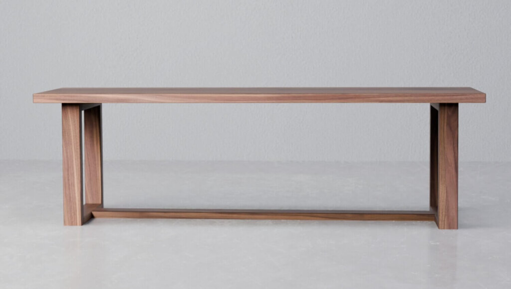 The Melissa Bench, a piece of home furniture, showcases a minimalist wooden design with a flat seat and rectangular legs. Positioned against a plain white wall on a light grey floor, it exudes contemporary aesthetics with its warm brown finish.