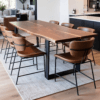 A modern dining room showcases a statement table with a live edge design and metal legs, surrounded by six brown chairs with black frames. The space features a light wooden floor and a white area rug, offering a glimpse into the kitchen and living area beyond.