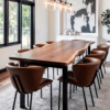 A dining room features a long live edge wooden table with black metal legs, surrounded by eight brown cushioned chairs. A modern light fixture hangs above, and a black and white abstract painting adorns the wall. Large windows flood the space with natural light.