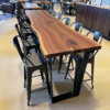 A spacious conference room boasts a long, sleek Black Walnut Single Slab Bar Table With U-Shaped Legs, showcasing smooth edges and a rich grain pattern. Black metal chairs are neatly arranged on both sides, with a counter featuring a coffee machine visible in the background.