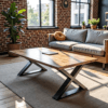A modern living room features The Summit Coffee Table with a sleek metal X-frame. Behind it, a gray sofa with yellow and gray cushions leans against a brick wall, while natural light floods in through large windows.