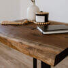 Close-up of The Black Walnut Live Edge End Table with a natural rustic finish, featuring a notebook, a "Leather" candle, a wooden bead garland, and a textured white vase against a plain background.