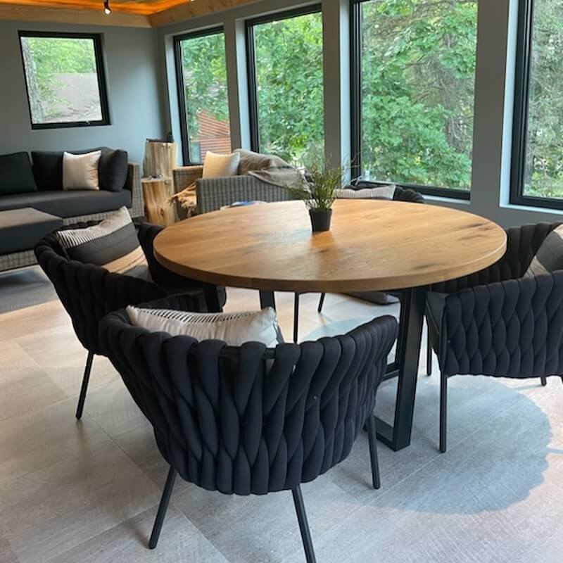 Cozy room with a round Fargo Woodworks dining room table surrounded by four black woven chairs. A small plant sits gracefully on the table, enhancing the dining experience. Large windows reveal greenery outside, while a cushioned bench invites relaxation in the corner.