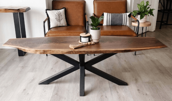 A modern living room showcases a wooden coffee table with a natural edge and black metal legs, embodying unique designs in home décor. Two brown leather chairs with pillows sit in the background, accompanied by potted plants on side tables. The floor is light-colored wood.