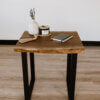The Black Walnut Live Edge End Table, featuring elegant black metal legs, stands on a light wood floor. It holds a small vase with flowers, a candle, a decorative tassel with beads, and a closed notebook topped with a pen.