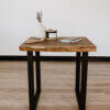 The Black Walnut Live Edge End Table has black metal legs and rests on a light wooden floor. Its surface holds a white vase with lavender, a candle, a notebook, and wooden beads. The backdrop is a plain white wall.
