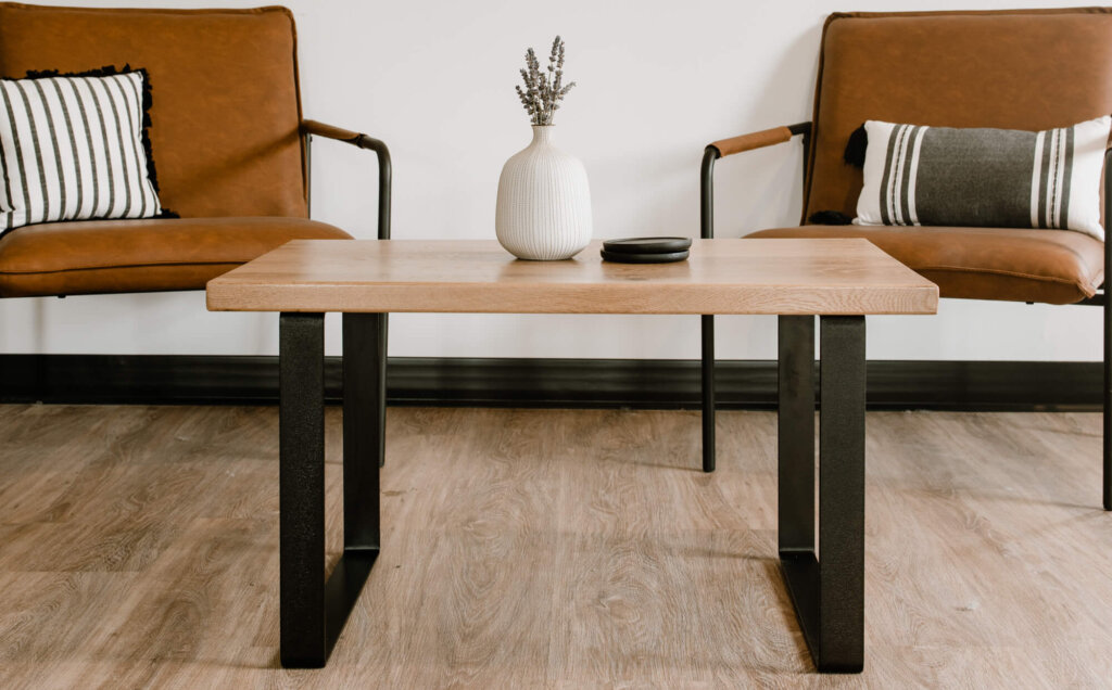 A minimalist living room features two stylish brown chairs, each adorned with a striped pillow, flanking a durable white oak coffee table. The table holds a small white vase with lavender. The room's light wood flooring complements the plain white wall effortlessly.