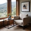 A cozy beige armchair is positioned by a window with a scenic forest view. The Sedona End Table nearby holds a vase with greenery and a book. A framed black and white artwork adorns the wall, while a light rug covers the wooden floor.